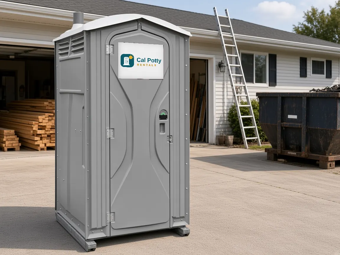 A grey porta potty on a driveway at a home remodel with lumber and a dumpster, an example setup for porta potty rental in East Palo Alto