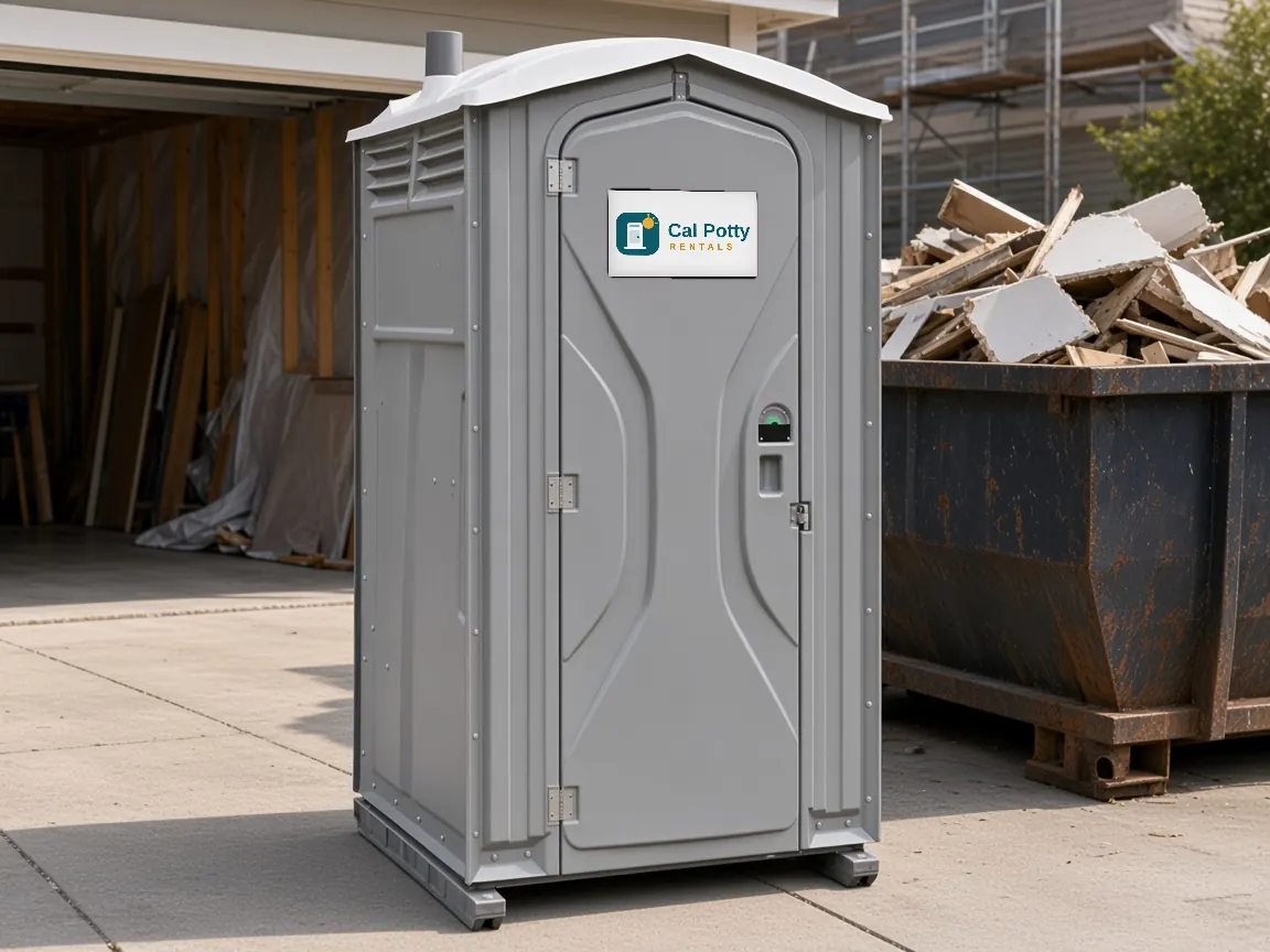 A grey standard porta potty beside a driveway garage remodel with a debris dumpster